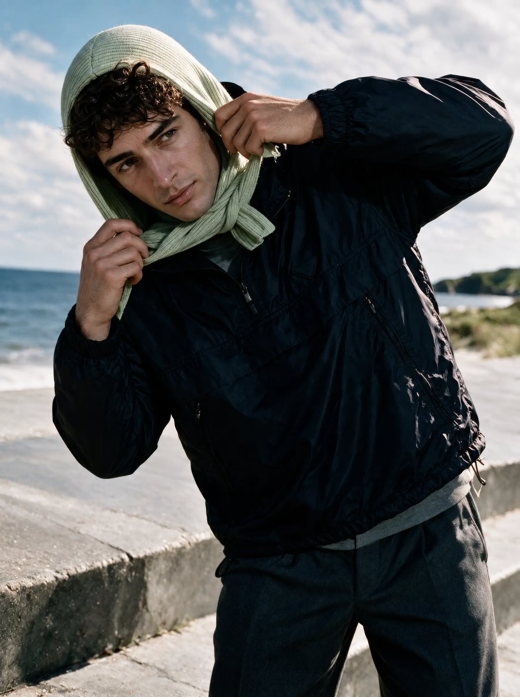 A model in a dark jacket pulling up a pale hood by the sea